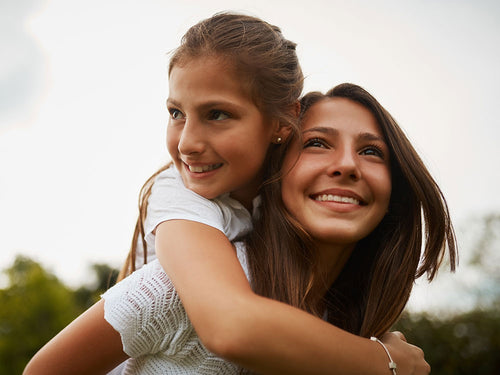 A teenage girl gives another a piggy back, looking to the sky busting through the blocks. 